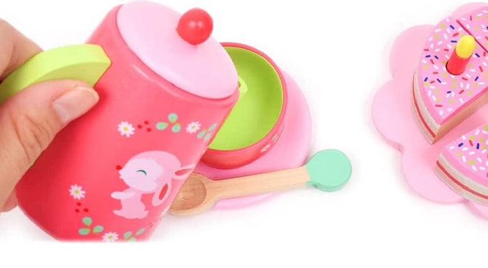 Pink wooden tea pot with a lime green handle, pink and green tea cup, wooden spoon, and pink cake on a white background