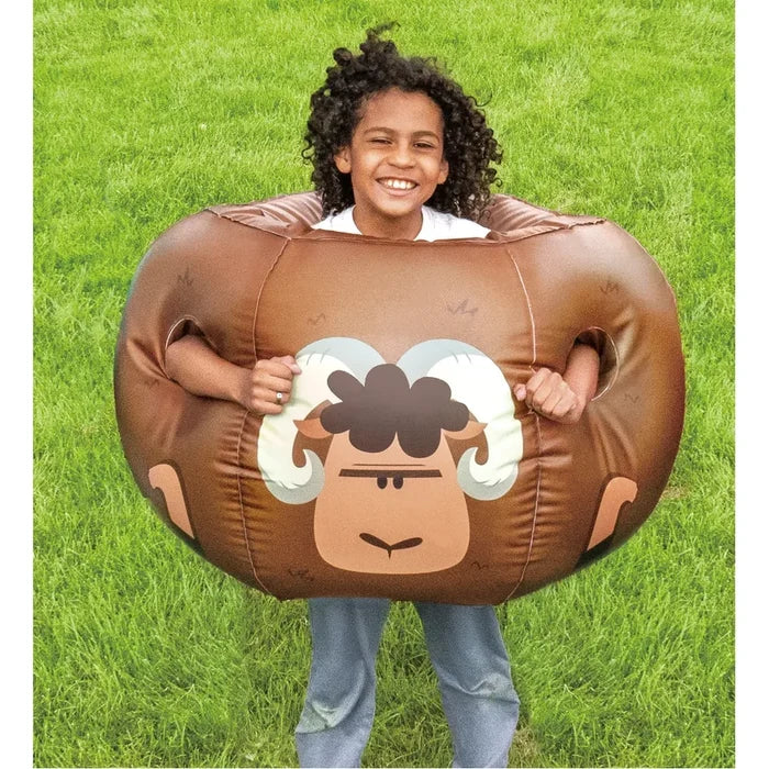 A child wearing a brown inflatable ram bumper ball on a green grass lawn. The ball features a printed ram face with white and dark brown horns and a simple black mouth.