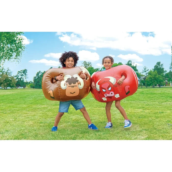 Two children outdoors on a grassy park lawn wearing inflatable bumper balls, one brown and one red, under a blue sky with clouds.