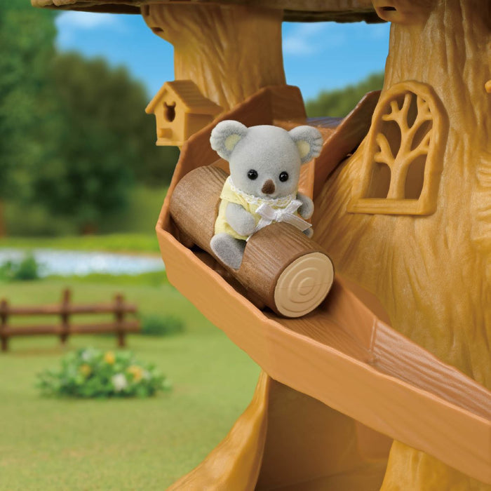 A small gray koala figurine wearing a yellow dress sits on a wooden log inside a treehouse slide, with a blurred outdoor garden background.