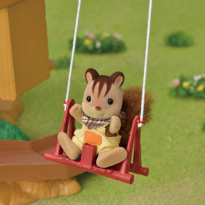 A small plush squirrel toy wearing a yellow outfit and plaid scarf, sitting on a red swing in a garden-themed setting with green grass and a wooden structure blurred in the background.