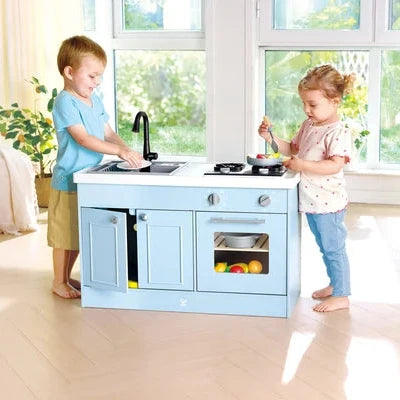 Two children playing with a light blue wooden toy kitchen island in a bright, sunlit room. One child stands at the sink on the left, and the other stands at the counter on the right.