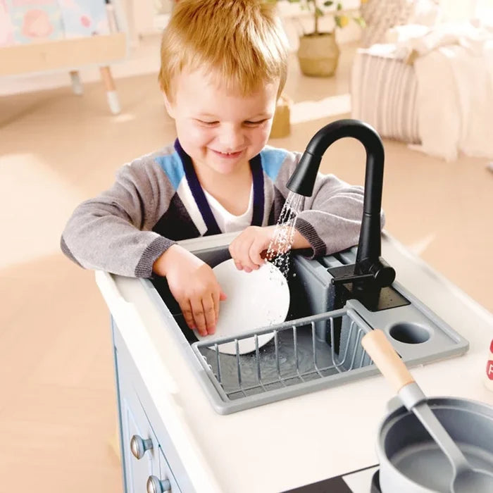 A young child smiling while washing a white plate at a toy kitchen sink with a black faucet and gray drying rack, in a bright indoor room.