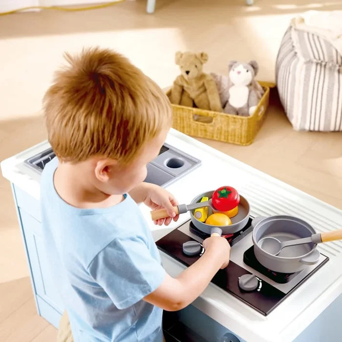 A young child in a light blue t-shirt plays with a toy kitchen stove, cooking pretend food in gray pans. The scene is set in a brightly lit room with wooden floors, a striped pouf, and a basket of stuffed toys in the background.