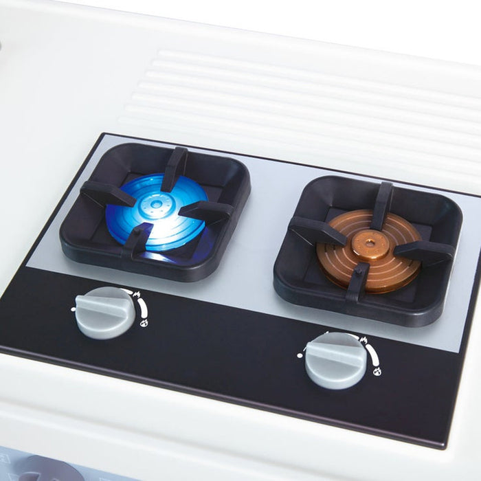 A close-up of a pretend two-burner gas stove top with black grate burners, one lit with a blue flame and the other with a brown flame, set into a white stovetop cooktop with two silver control knobs.