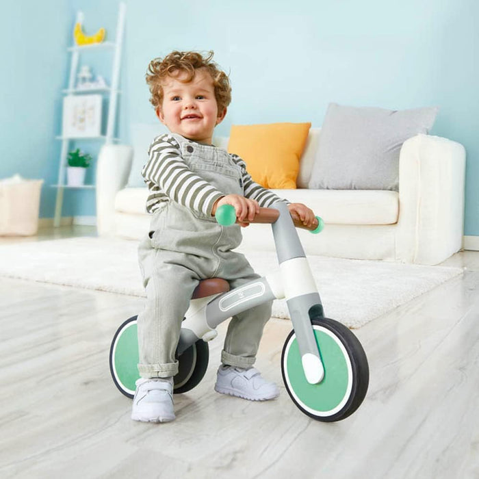 A young child sitting on a green and white Hape First Ride Balance Bike indoors.