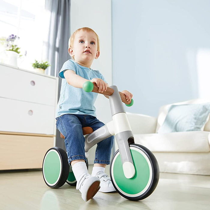 Child riding a green and white balance bike in a room with light-colored furniture.
