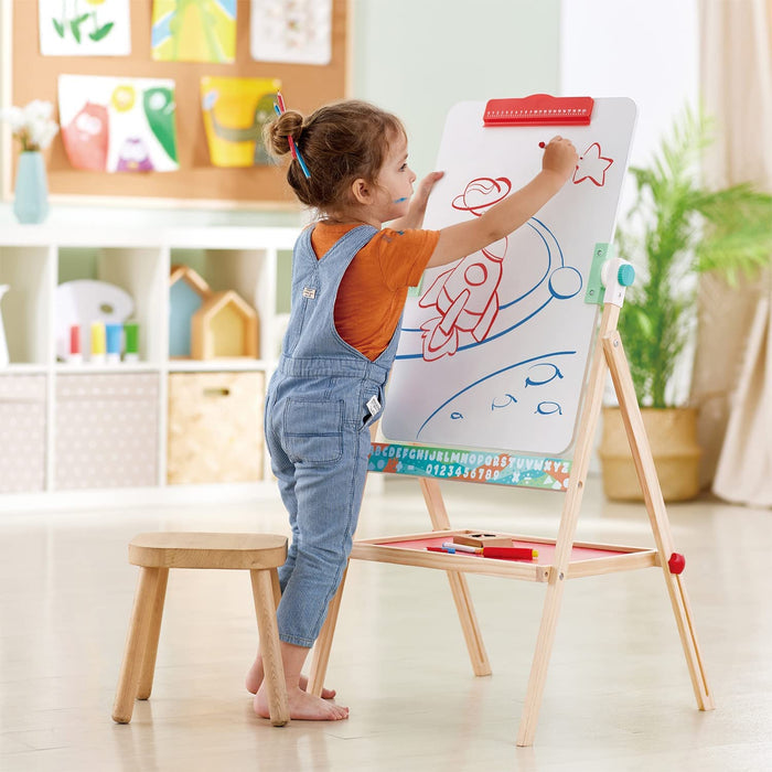 Child drawing on a whiteboard easel with colorful markers in a bright room.