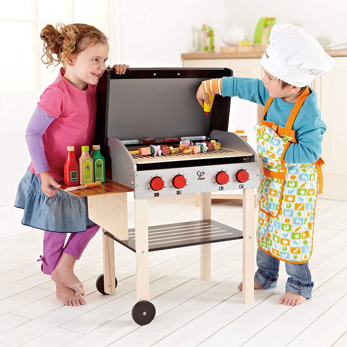 Two children, one wearing an apron and chefs hat, playing with the Gourmet Grill in a playroom.
