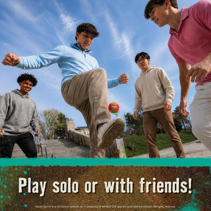 Four young men outdoors playing with a small orange Hacy Sack under a blue sky. One man in a light blue hoodie kicks the ball while the others watch.