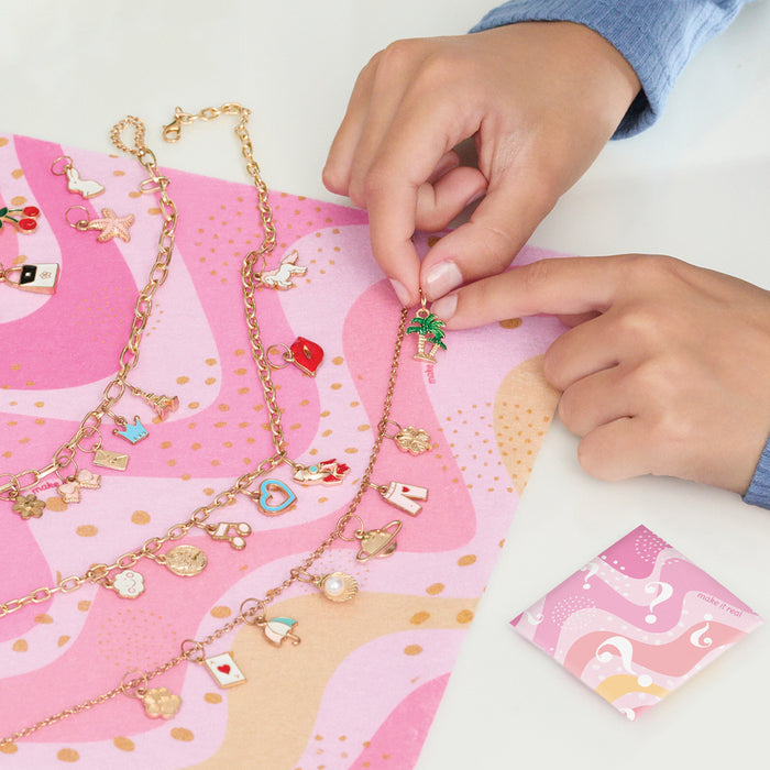 Two hands attaching a charm to a chain on a white table, with a colorful pink and gold spotted table mat on the table