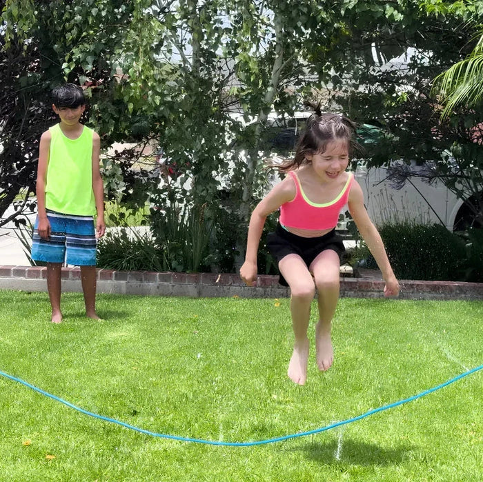 A young girl in a pink and black swimsuit jumps barefoot over a blue water hose in a green backyard, with a boy in a neon yellow shirt standing in the background.