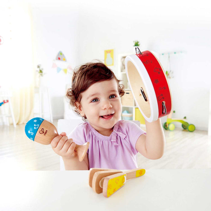 A young child with curly brown hair smiles while holding wooden toy instruments in a bright, softly lit room. The child is wearing a light pink top and holds a maraca with a blue top and a tambourine with a red top.