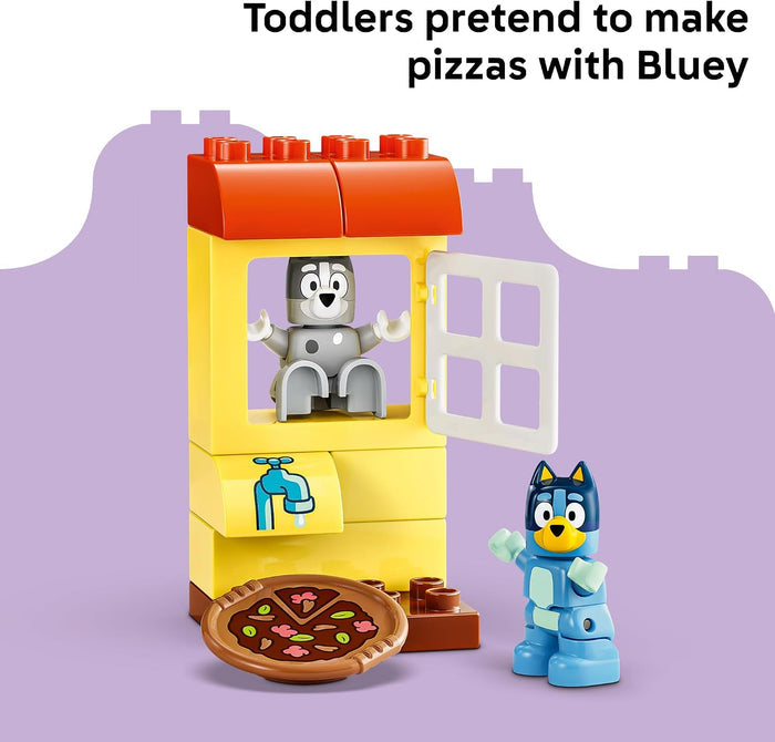 A children's toy set showing a yellow and orange pretend kitchen scene with a gray robot inside and a blue dog figure beside it, plus a pizza piece on the floor. Text at the top reads 'Toddlers pretend to make pizzas with Bluey'.