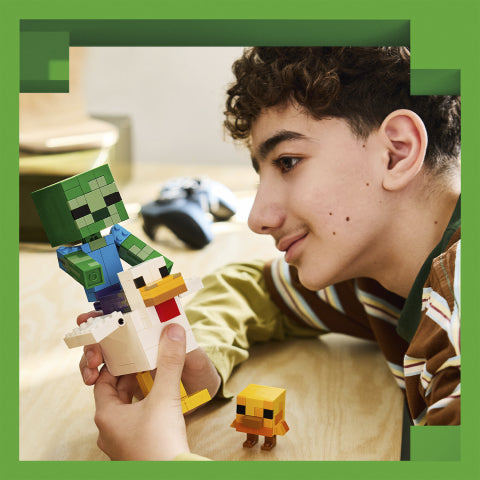 A young boy with curly brown hair smiles while looking at a hand holding a small Minecraft-style chicken figure. Another small blocky character figure sits on the table nearby, with a blurred game controller in the background.