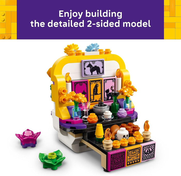 A colorful toy building-block set displayed on a white background with a purple banner at the top reading 'Enjoy building the detailed 2-sided model.' The set includes a decorative altar-like structure in yellow, purple, black, white, orange, and pink, with small accessories such as flowers, cups, and figurines.