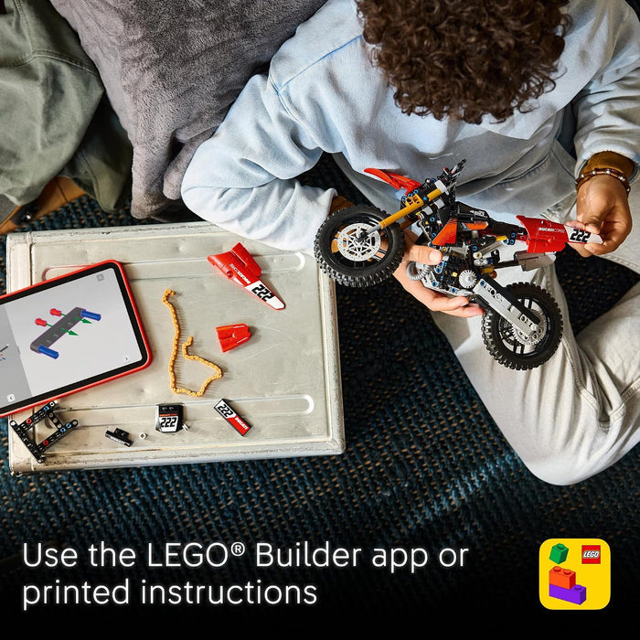 Top-down view of a person building a red and black LEGO dirt bike using loose bricks on a tray, with a tablet showing the LEGO Builder app and printed instructions. Text at the bottom says, 'Use the LEGO Builder app or printed instructions.