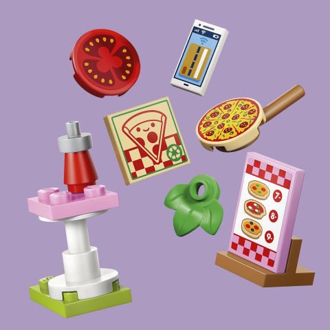 A set of toy construction blocks arranged on a solid lavender background, including a pizza oven, smartphone, pizza pan, pizza slice, green pepper slice, and a pink-and-white menu board with pizza graphics.