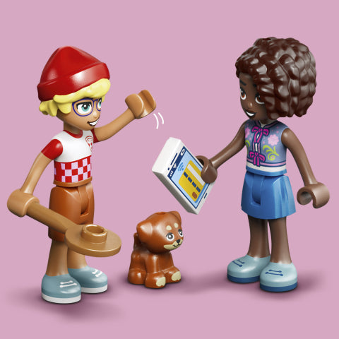 A LEGO Friends toy set featuring two female minifigures and a small brown dog on a solid pink background. One figure wears a red and white checkered shirt, brown pants, a red beanie, and blue shoes, holding a plate.