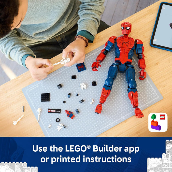 A person is assembling a LEGO Spider-Man figure on a gray grid mat over a wooden table, with loose building blocks around the figure and a tablet nearby. A LEGO app instruction graphic appears at the bottom.