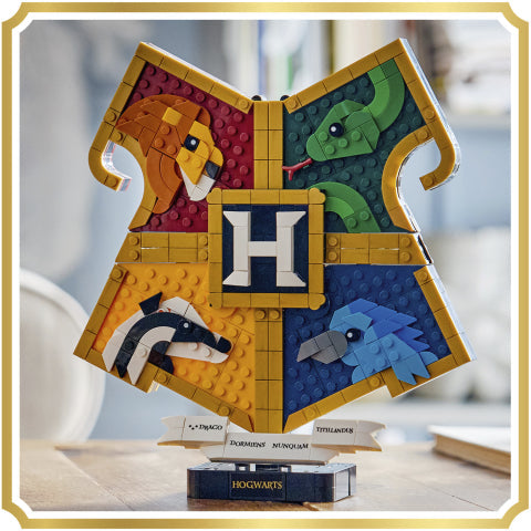A LEGO Harry Potter Hogwarts crest building set displayed on a small stand. The golden-yellow cross-shaped crest features four colored quadrants with the lion, snake, badger, and raven, surrounding a central navy blue square with a white letter H.