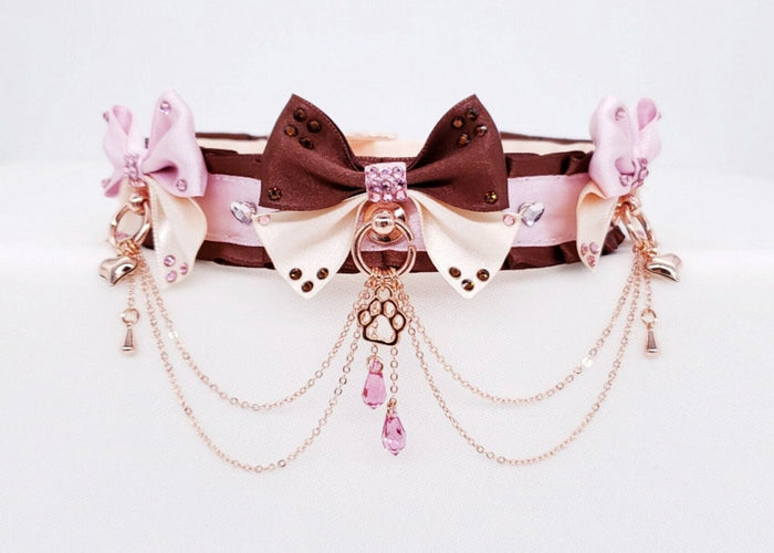 A deluxe pet play collar in chocolate, pink, and cream colors, featuring satin and nylon materials, decorated with a two-tone center bow, small cream and pink bows, rose gold hardware, chains, and a rose gold paw charm.