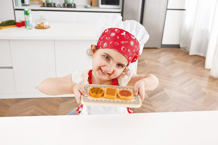 Child in a kitchen wearing a chef's hat and apron, holding a tray with colorful cookies.