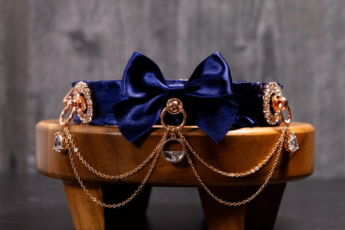 An ornate navy and rose gold collar with a large navy bow, rose gold O-rings, chain work, and crystal embellishments displayed on a wooden stool.