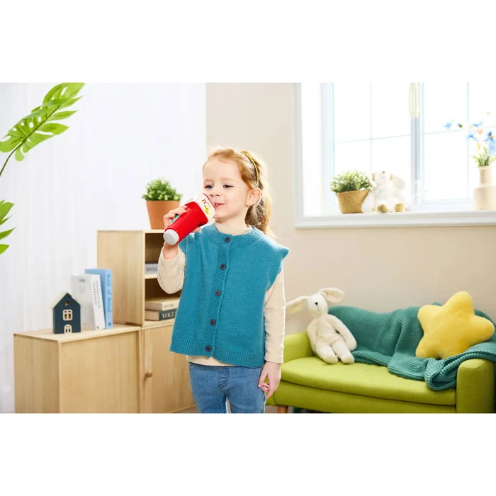 A young girl stands in a bright, modern children's room wearing a sleeveless teal knit vest over a cream long-sleeve top and blue jeans. She is holding a red cup near her mouth.