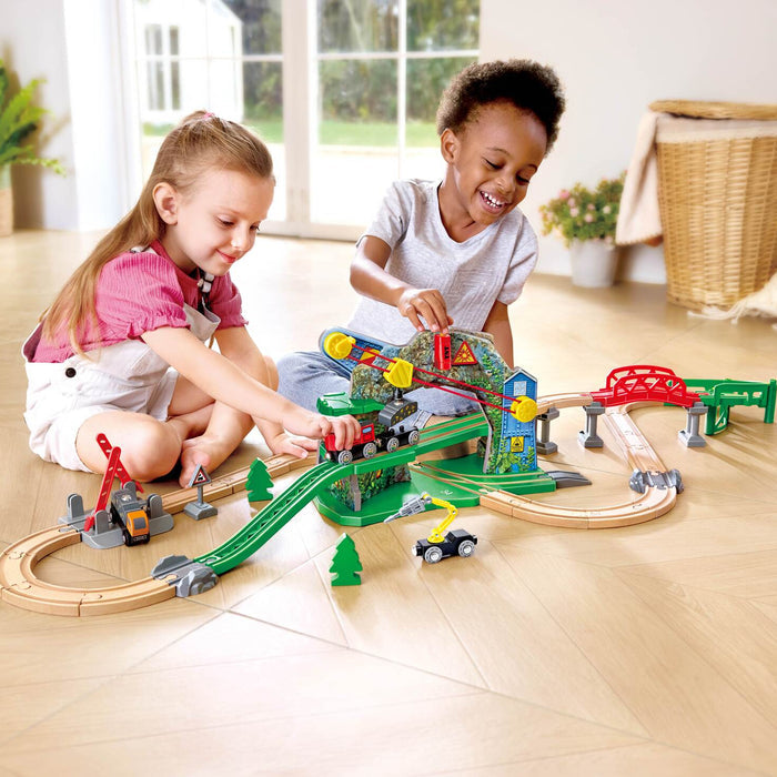 Two children playing with a colorful wooden toy train set on a light wood floor in a bright room. The toy set includes curved tracks, a green bridge, a red bridge, a mountain pass, and small train cars.