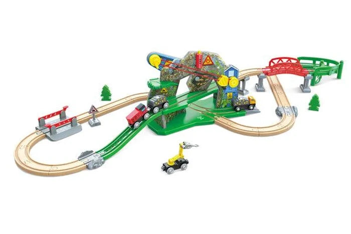 A children's wooden toy train set with curved and straight wooden tracks, green tracks, a small bridge, a ramp, a dinosaur-themed station, and several toy trains and accessories, shown on a plain white background.