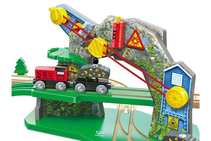 A wooden toy train bridge and signal system set on a white background. The green wooden track features a curved bridge with a blue signal building, a gray rock face with yellow pulleys, and a red and black toy train.