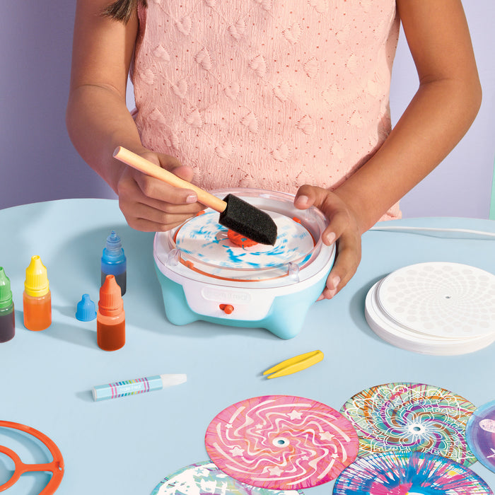 A child wearing a pink textured top uses a small light blue and white spin art paint splatter machine on a light blue table. The scene includes colorful paint bottles, a sponge brush, a white lid, and several round patterned paper discs.
