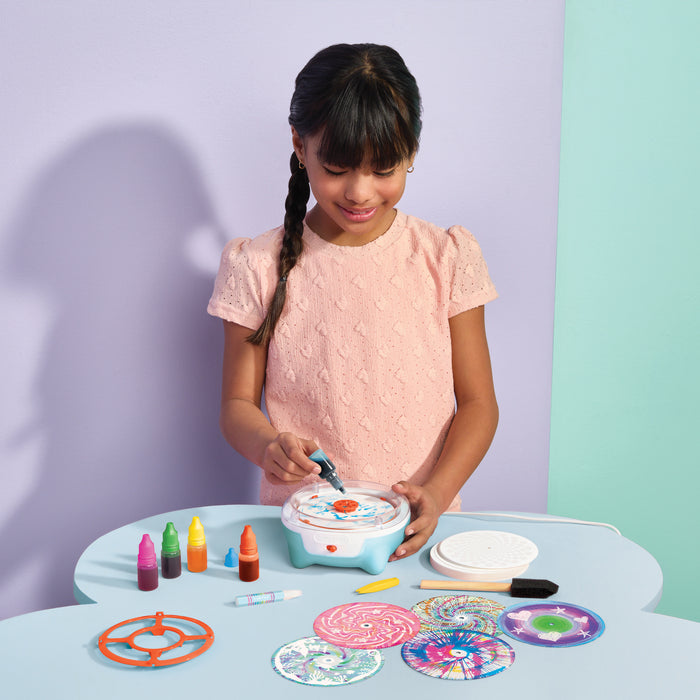 A young girl with braided hair smiles while using Utlimate Spin Art on a light blue table. The toy has a white and light blue body with colorful circular components, surrounded by paint bottles, circular discs, and tools.