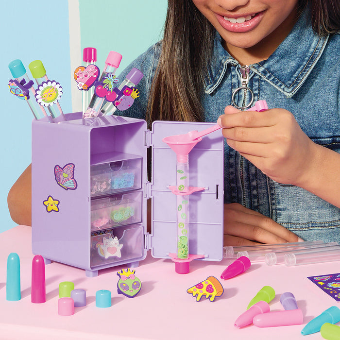 A child's craft locker shown on a pink table with a smiling girl partially visible. The lavender locker has clear drawers, stickers, colorful makeup pens, and a syringe-style applicator, with additional accessories scattered around.