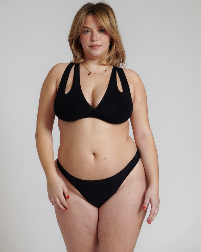 A person wearing a black x back system and black thong on a plain background.