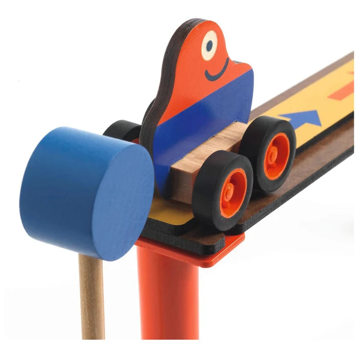A close-up of a wooden toy train set showing a small red and blue train car with black wheels on a brown track, alongside a blue cylindrical piece and orange support, on a white background.